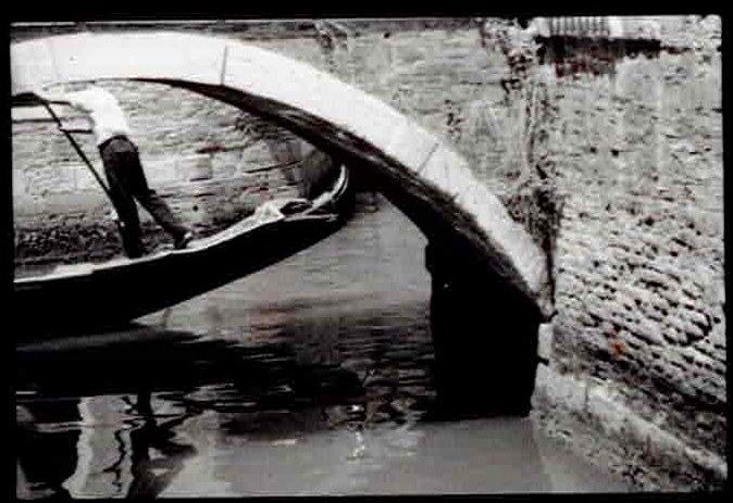 Black and white film photograph of a gondolier passing beneath a stone bridge in Venice