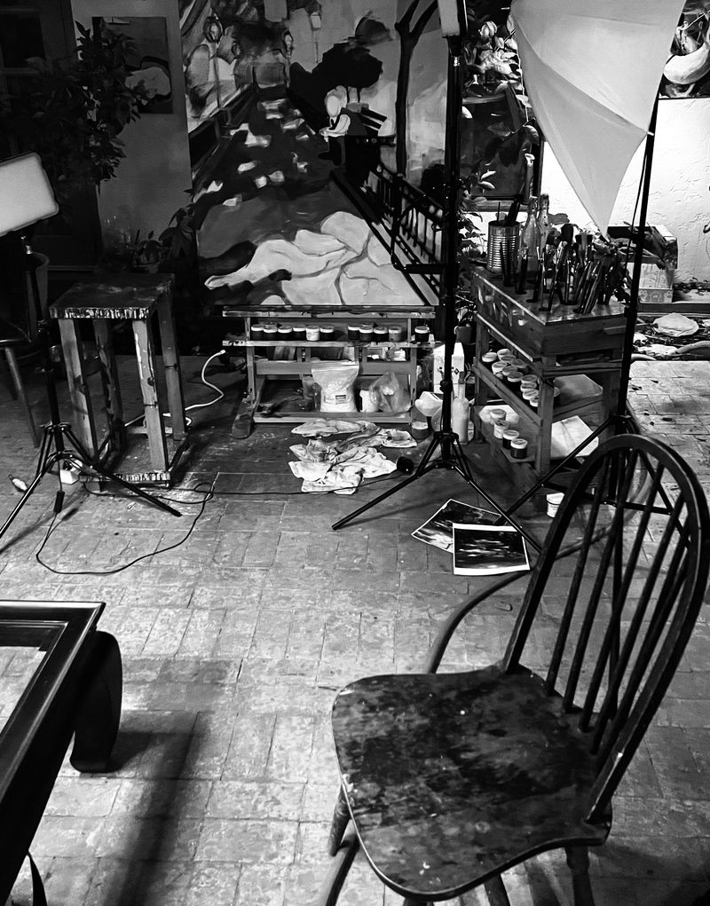 The studio at night in black and white: jars of pigment, brushes, rags on the floor, and a large painting in progress behind a wooden chair